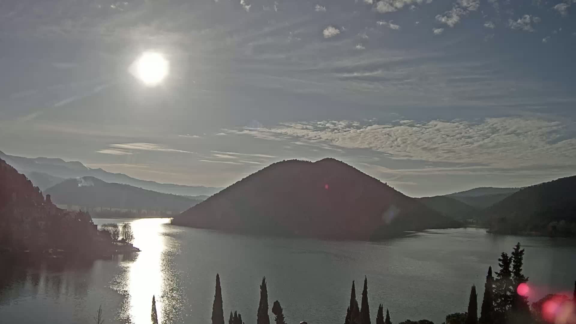 A bright sun shines over a calm lake nestled between low hills under a partly cloudy sky.