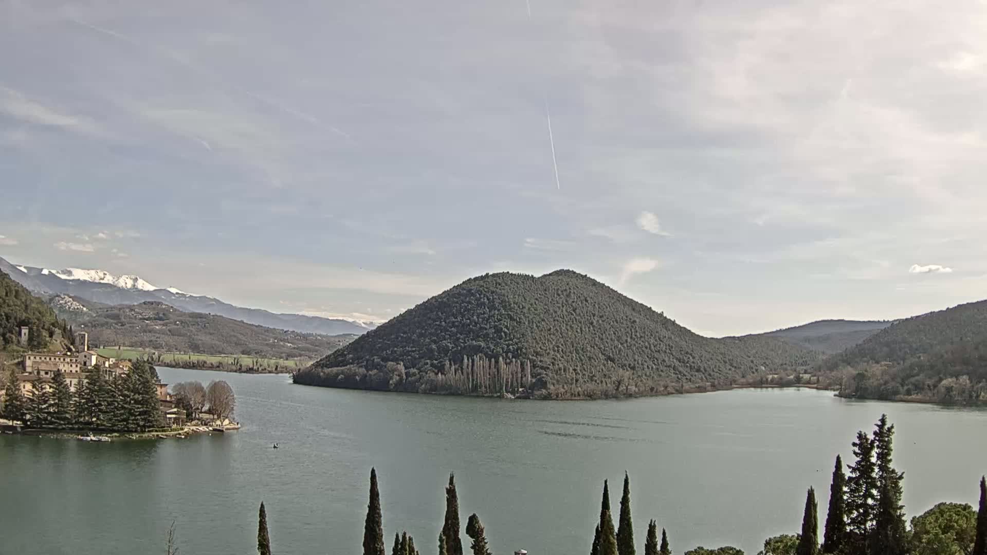 A lake with a small island and a village on the shoreline is nestled between rolling hills, with snow-capped mountains visible in the distance under a partly cloudy sky.