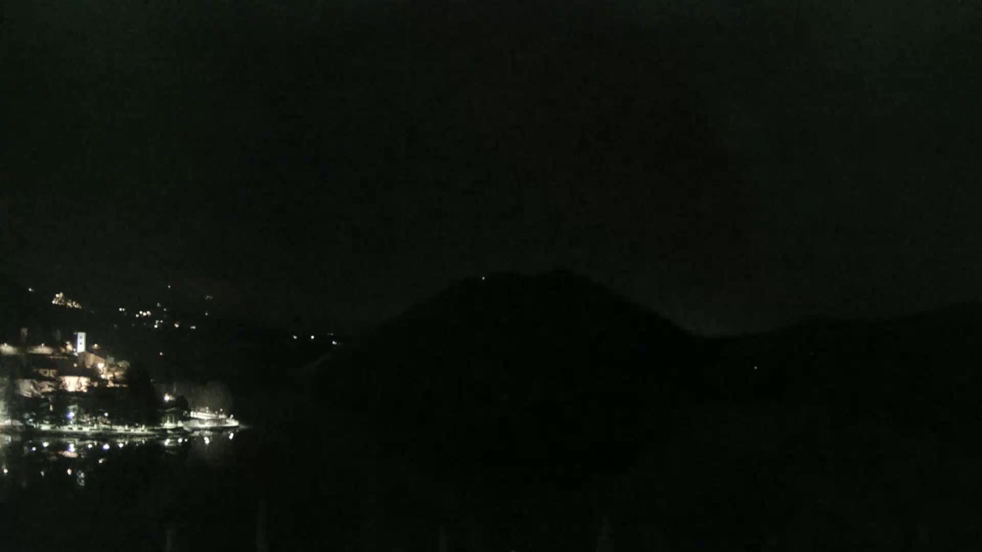 A dark, nighttime scene shows a body of water reflecting the lights of a town nestled on a hillside across from a large, dark hill.