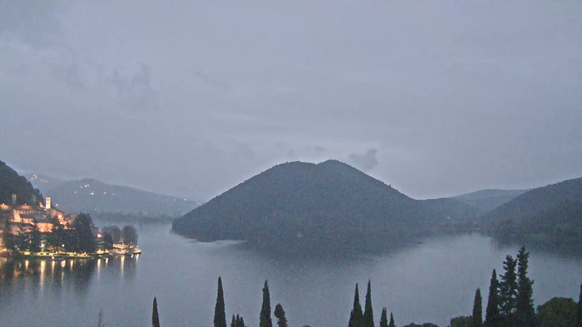 A lakeside town is illuminated at dusk under a hazy, overcast sky, with a large, dark hill rising from the water in the middle ground.