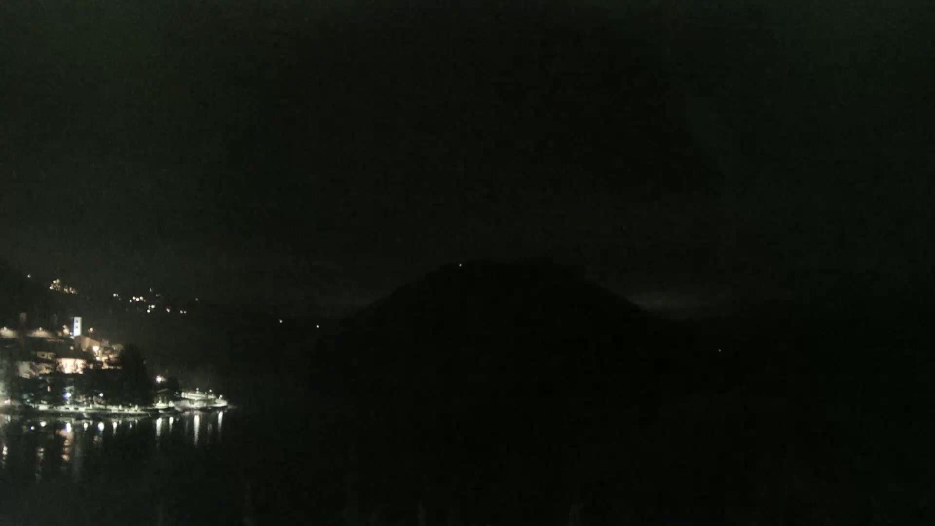 A nighttime view of a dimly lit lakeside town nestled between dark hills under a cloudy, dark sky.