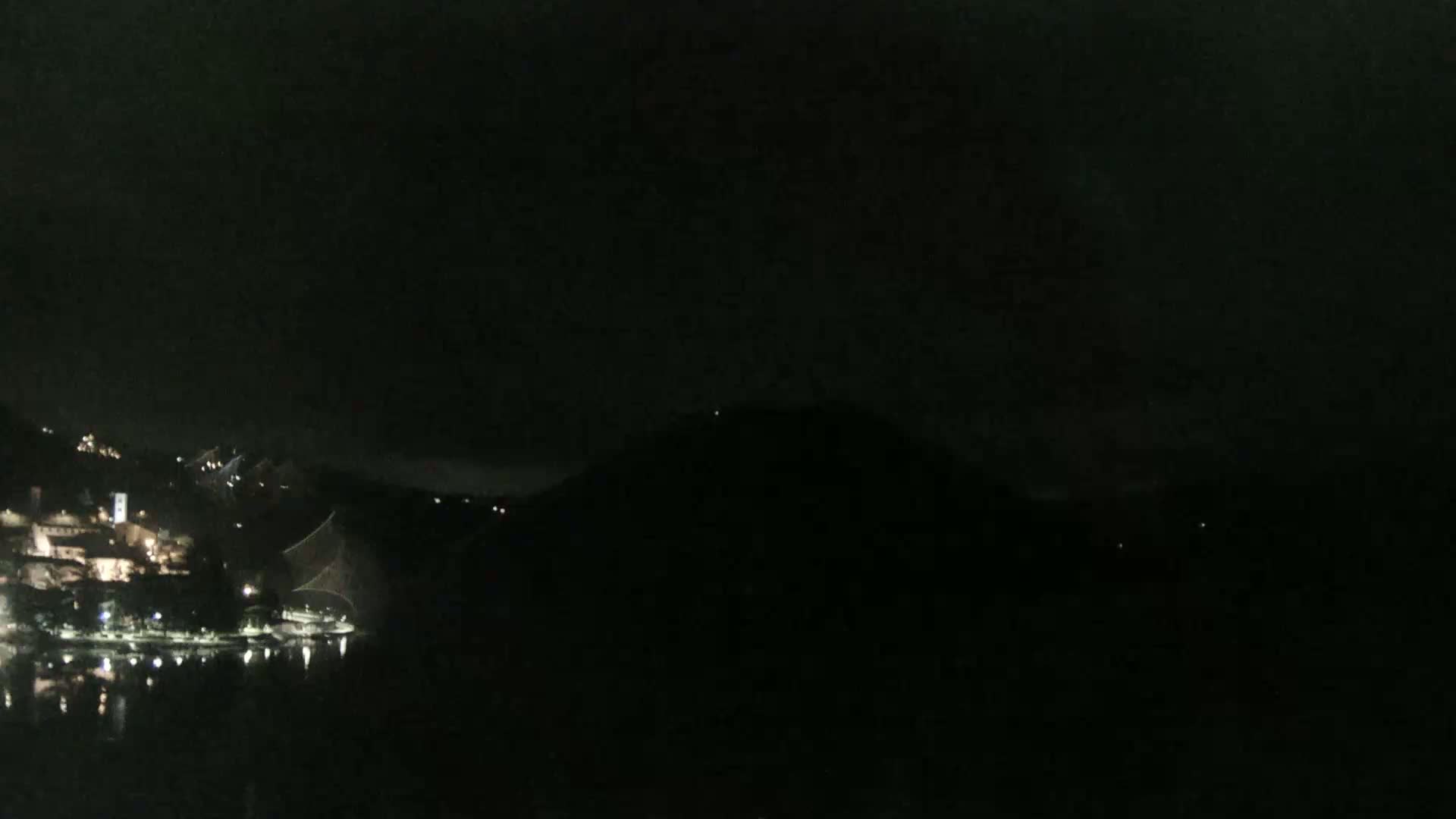 A nighttime view shows a dimly lit lakeside town nestled between dark hills under a mostly clear, dark sky.