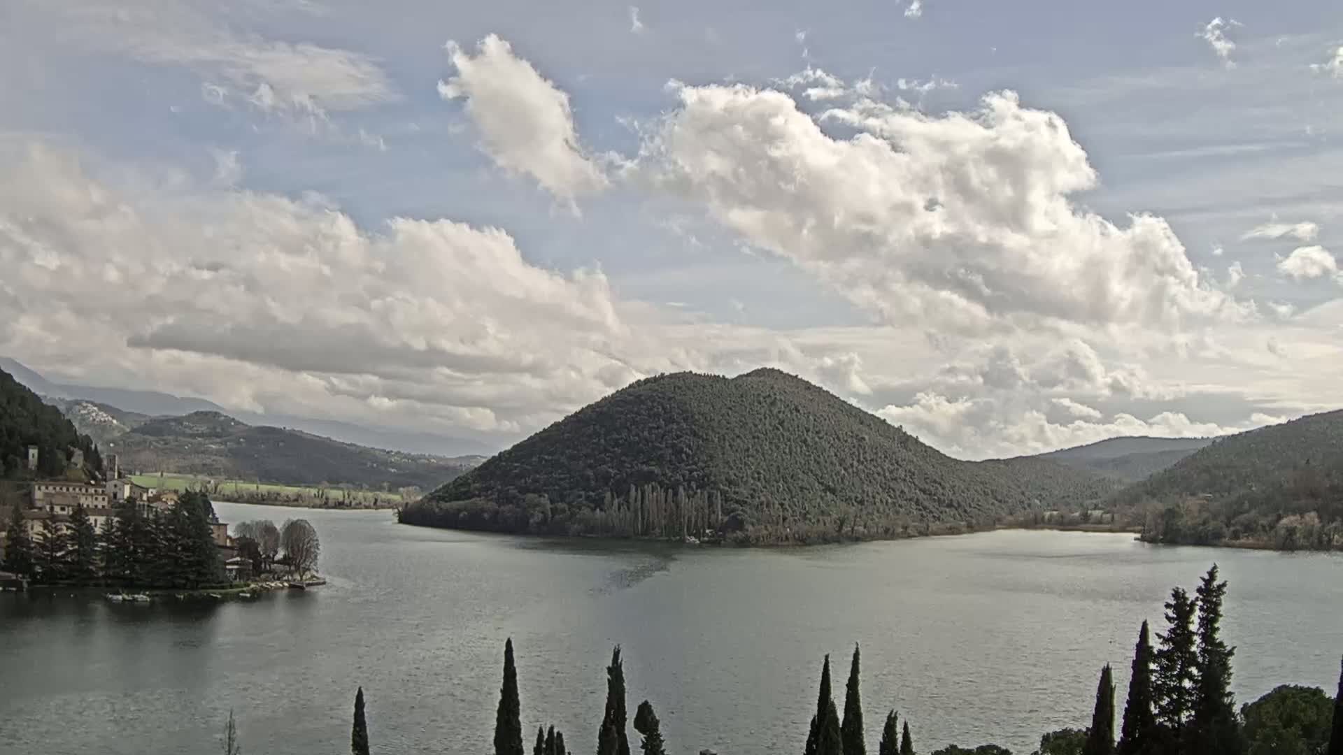 A lake is surrounded by hills and mountains under a partly cloudy sky, with a small town and island visible in the distance.
