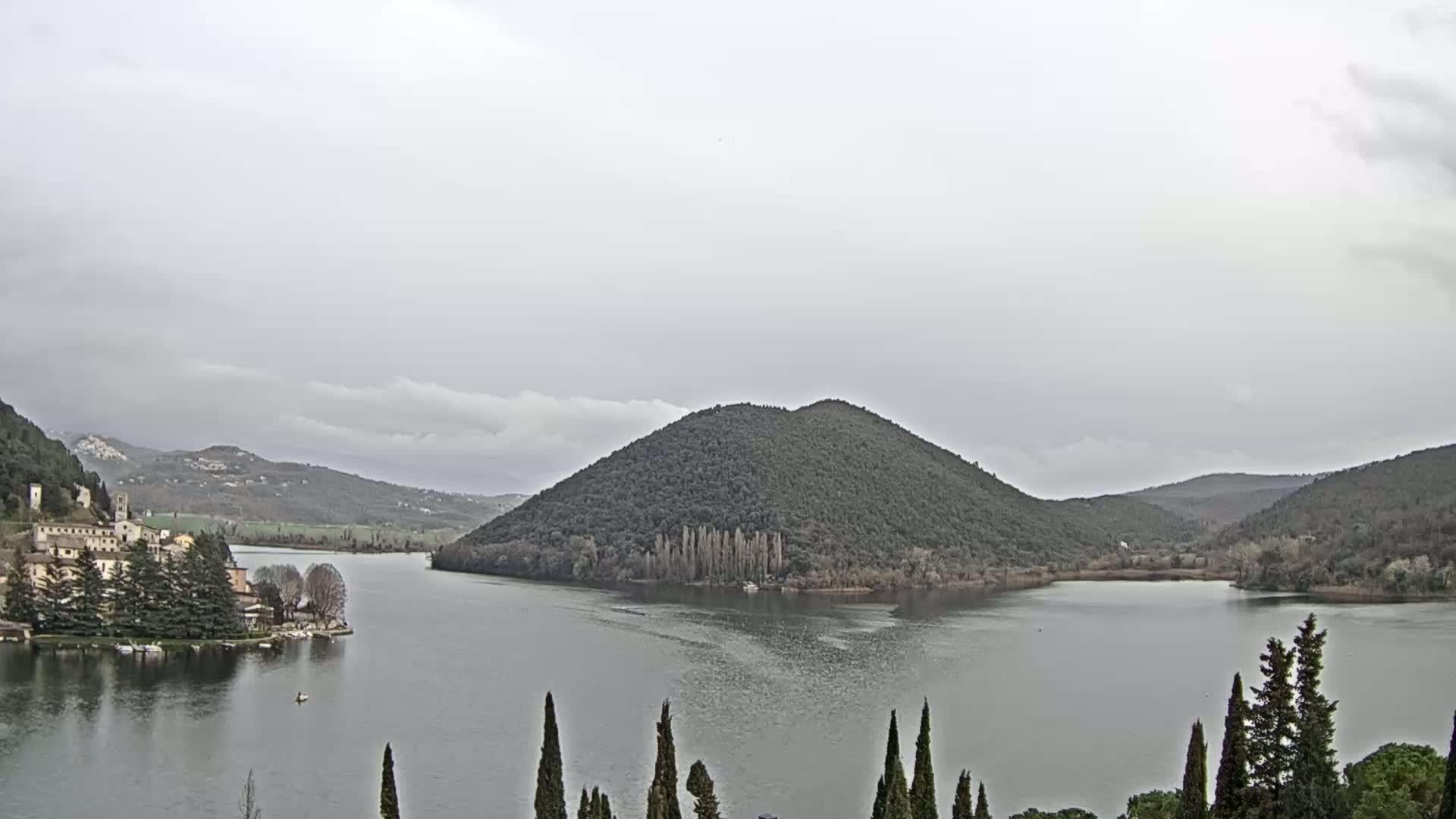 A lake is surrounded by hills and a small town, under an overcast sky.