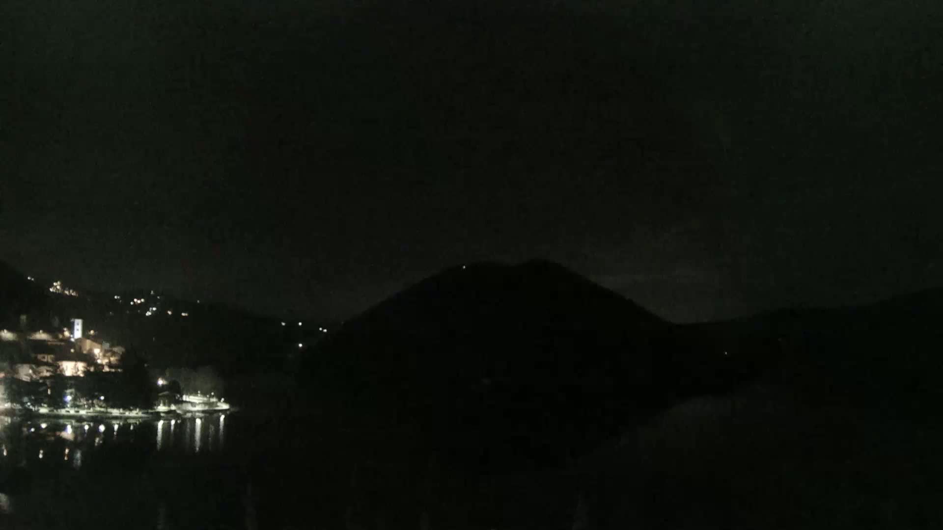 A dark night scene shows a body of water reflecting the lights of a town nestled in hills on one side, with a large dark hill silhouetted against the night sky on the other.