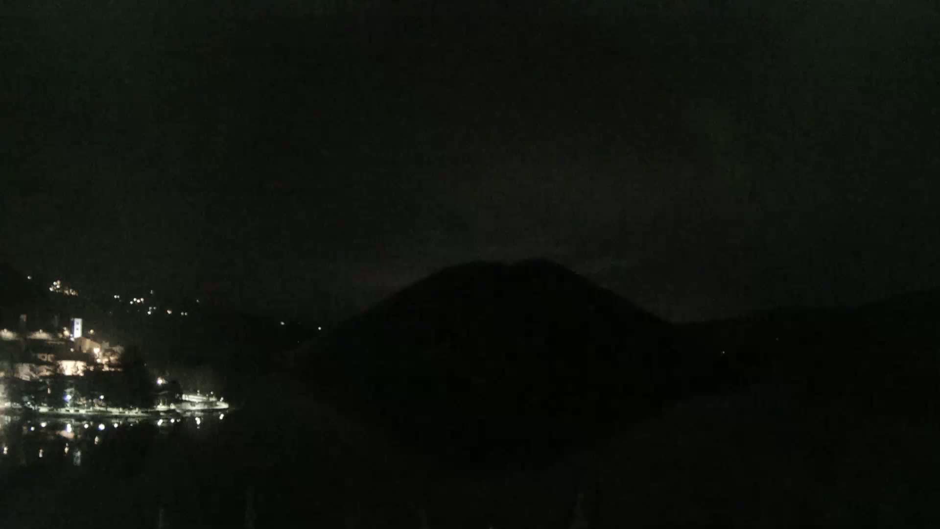 A dark, nighttime scene shows a body of water reflecting the faint lights of a town nestled in hills across the water, with a large, dark hill or mountain dominating the foreground under a mostly clear sky.