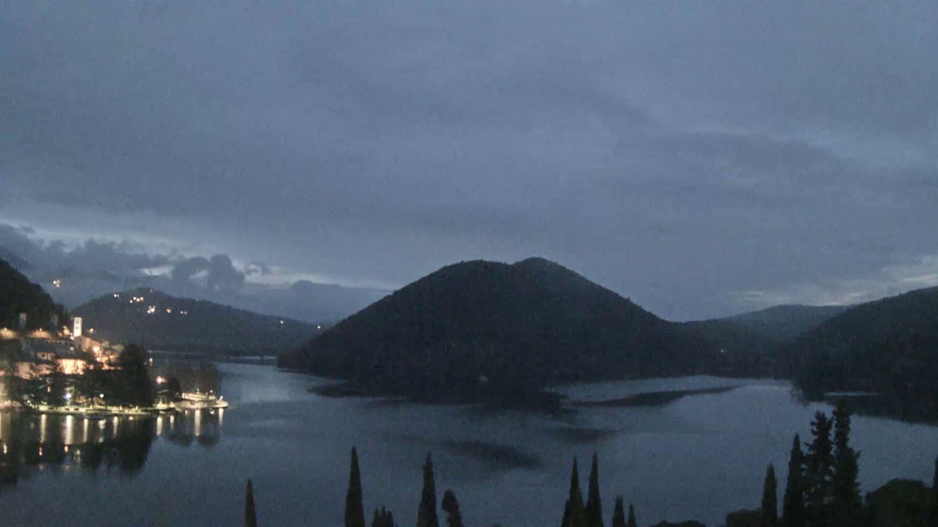 A dark, overcast evening scene shows a small town with lights on the shore of a calm lake, surrounded by dark hills and trees.