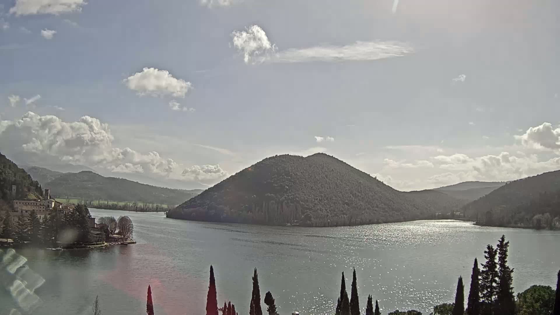 A partly cloudy day reveals a lake nestled between rolling hills, with a prominent, tree-covered island in the center.