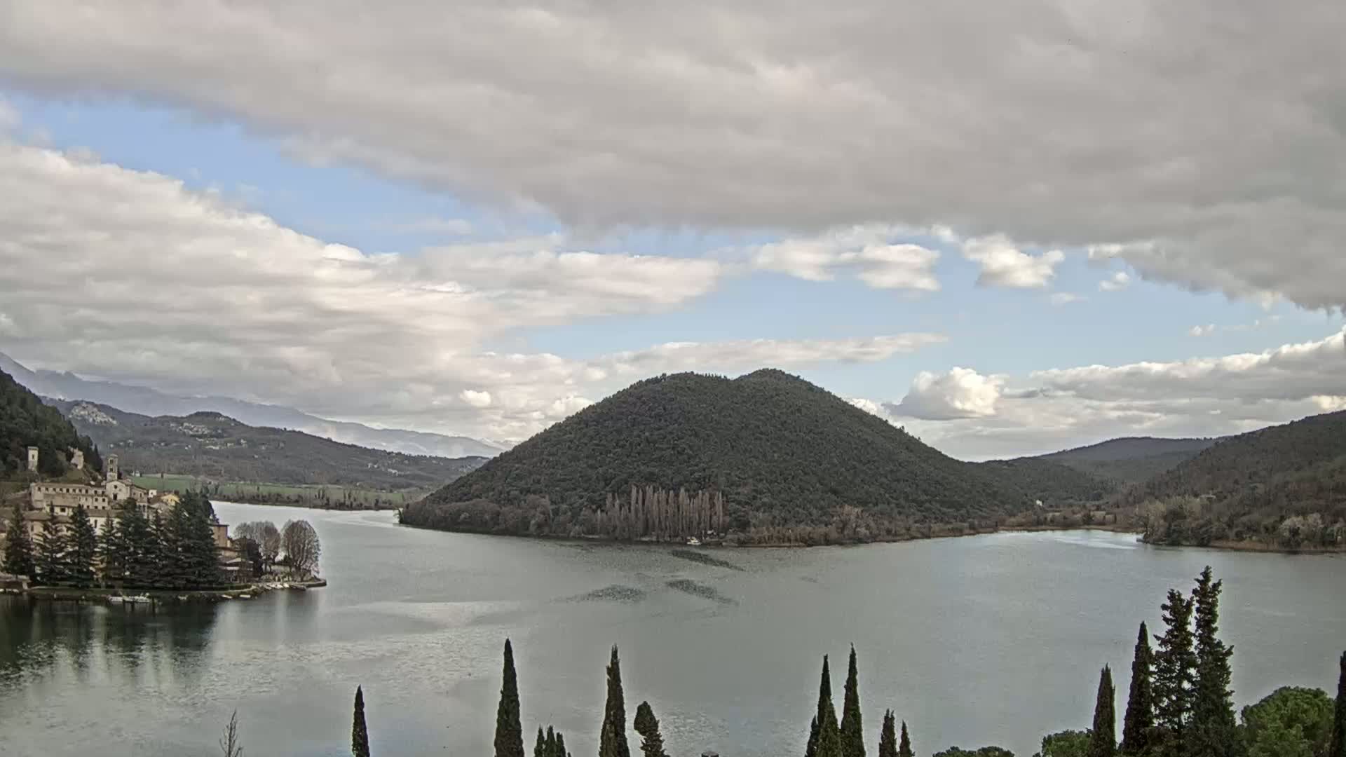 A lake is surrounded by rolling hills and a village, under a mostly cloudy sky.