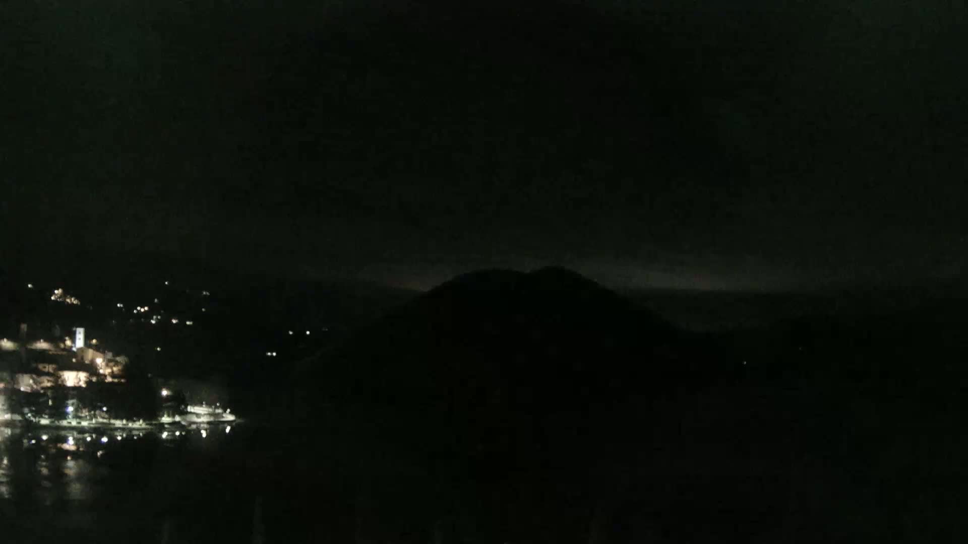 A nighttime view shows a dark hill silhouetted against a mostly clear, dark sky overlooking a dimly lit waterfront town.