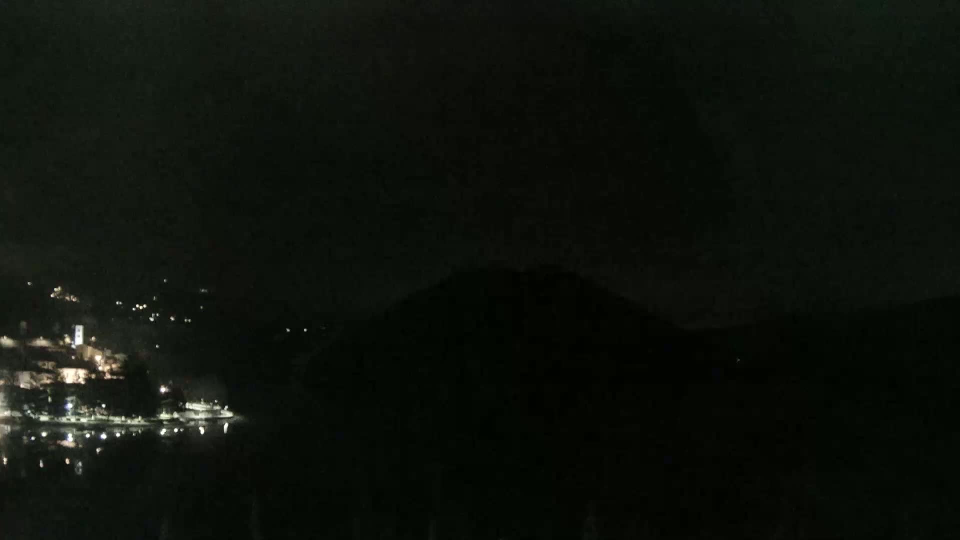 A dark, nighttime scene shows a body of water reflecting faint lights from a town nestled in hills on the opposite shore.