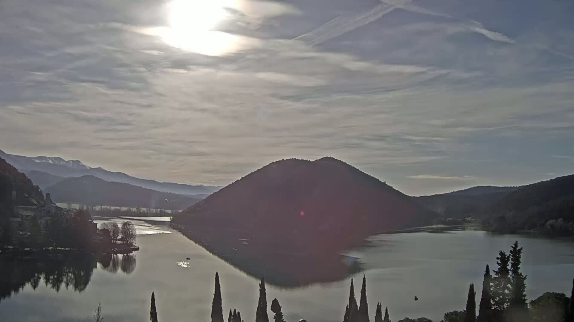 A calm lake reflects a sunlit sky and a dark, hill-shaped island, with distant mountains under a partly cloudy sky.