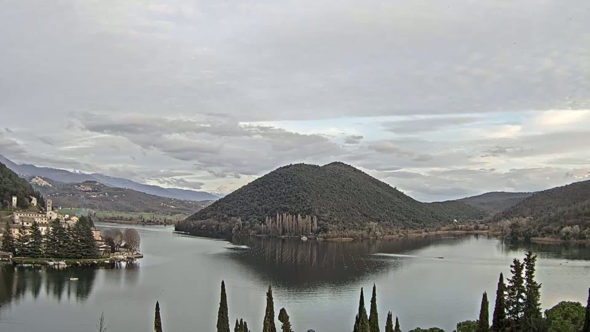 A calm lake reflects a tree-covered hill under a cloudy sky, with a village and mountains visible in the distance.
