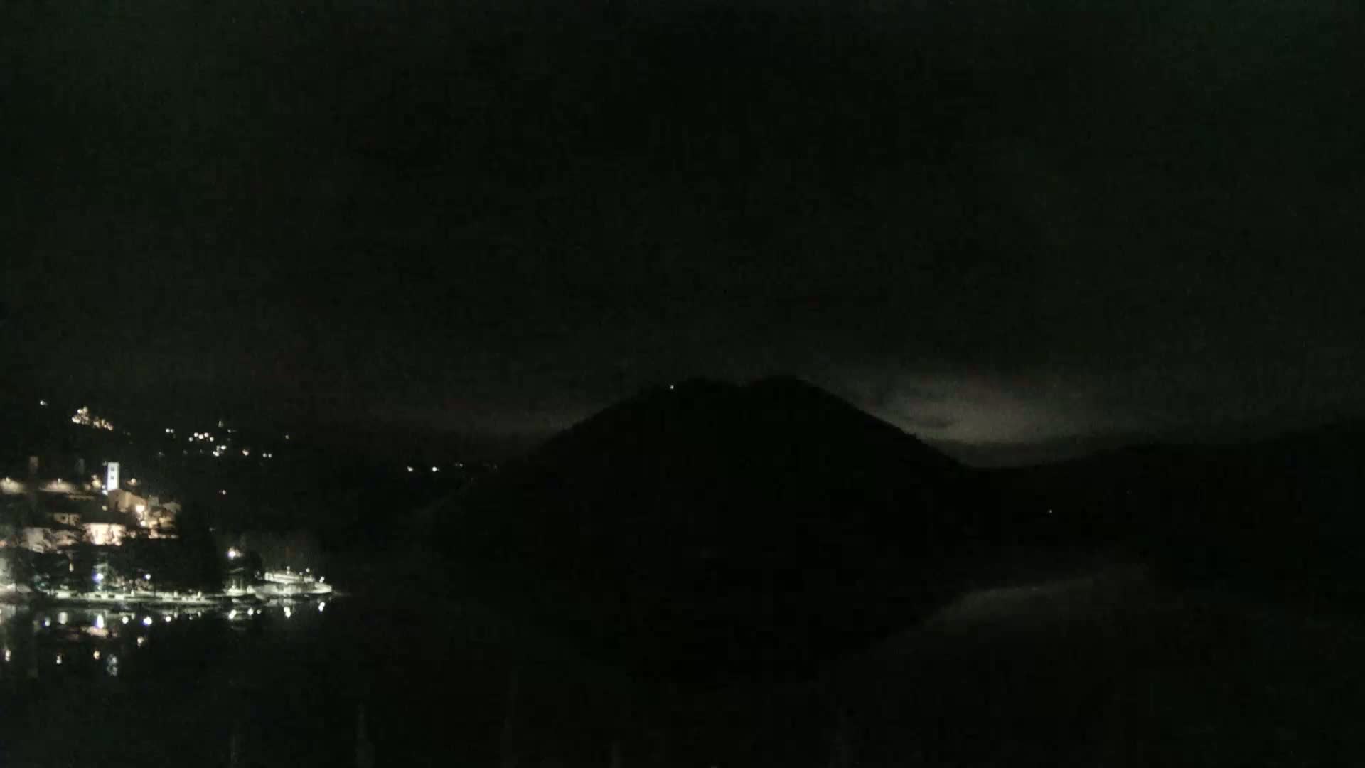 A dark, nighttime scene shows a body of water reflecting scattered lights from a village nestled at the base of a large, silhouetted hill under a cloudy sky.