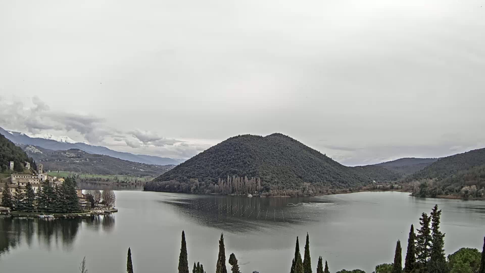 A calm lake is nestled in a valley between hills under a cloudy sky, with a village visible on the left shore and snow-capped mountains in the distance.