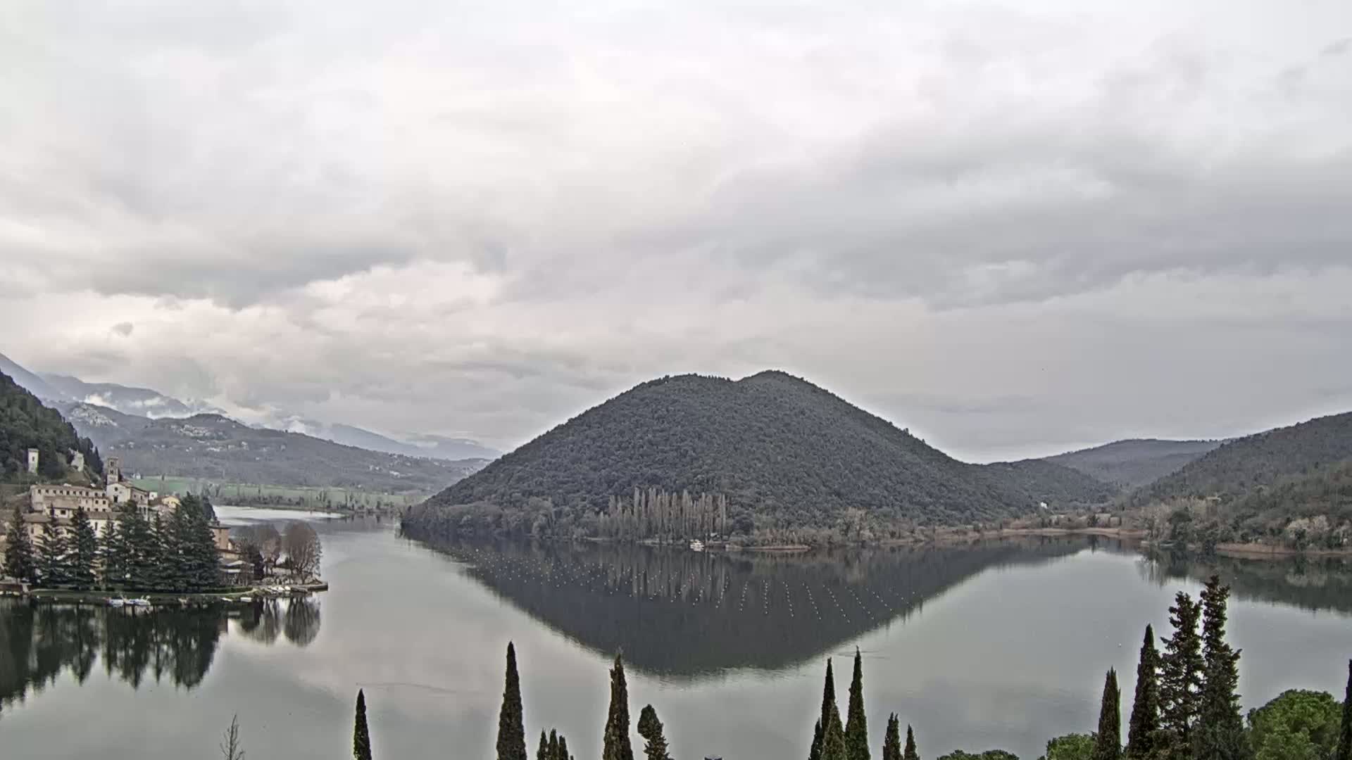 A calm lake reflects a tree-covered hill and distant mountains under an overcast sky.