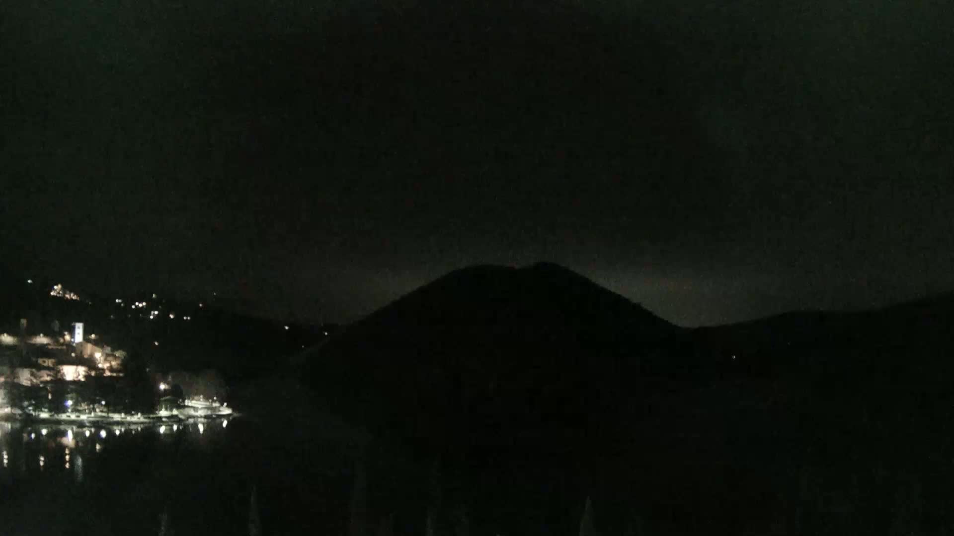 A dark, nighttime view shows a body of water reflecting a few lights from buildings on a hillside across the water, and a large, dark hill silhouetted against a mostly clear, dark sky.
