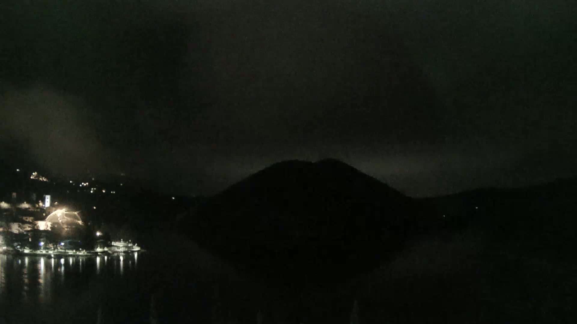 A dark, nighttime scene shows a body of water reflecting the lights of a town on a hillside, with a large, dark hill rising in the background under a cloudy sky.