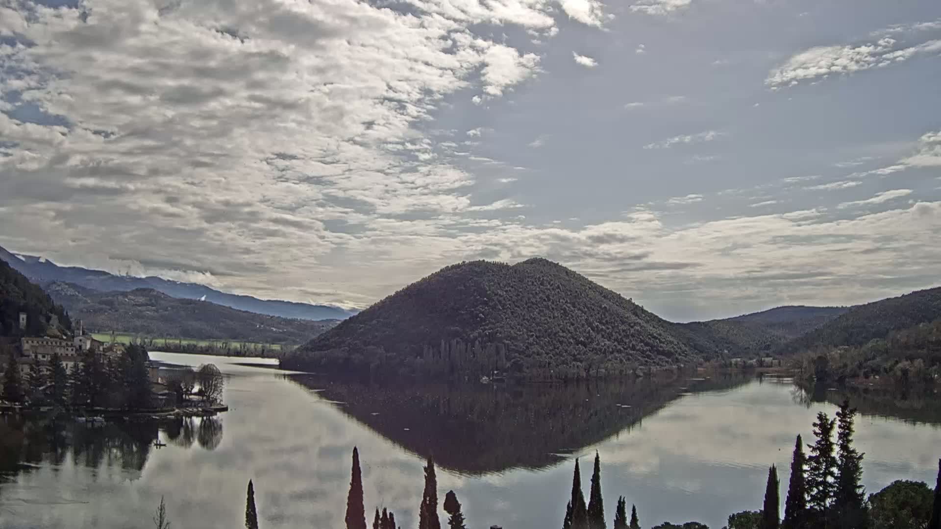 A calm lake reflects a tree-covered hill and distant mountains under a mostly cloudy sky.