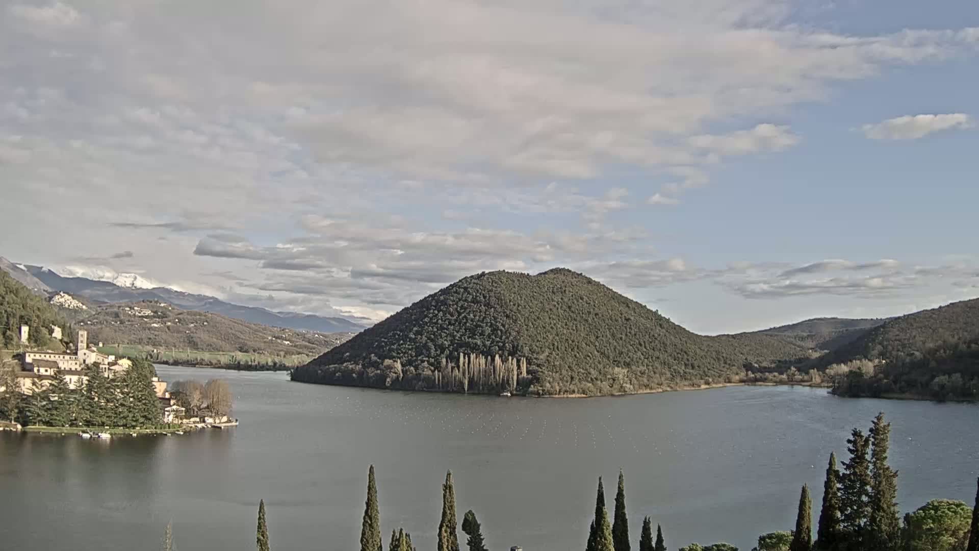 A lake is nestled between rolling hills under a partly cloudy sky, with snow-capped mountains in the distance.