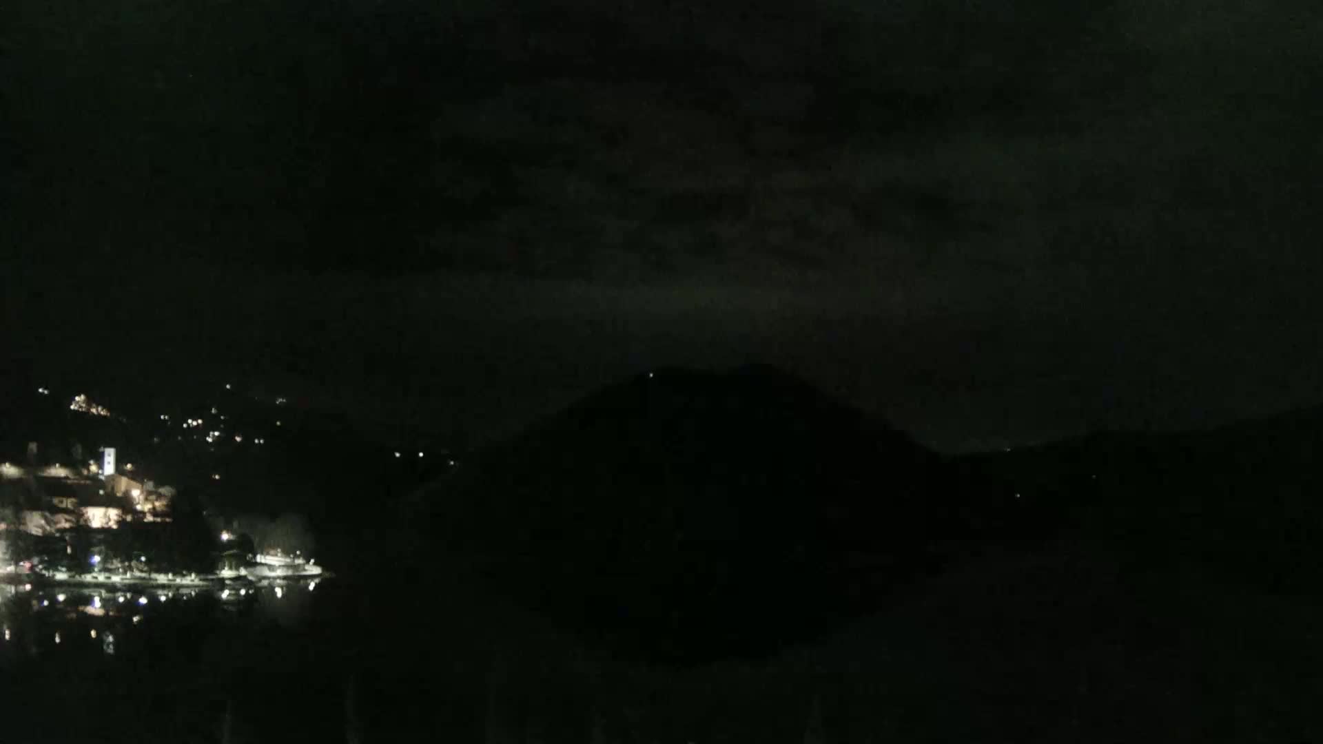 A nighttime view of a dark, hilly landscape with a body of water reflecting scattered lights from a town on the opposite shore under a cloudy sky.