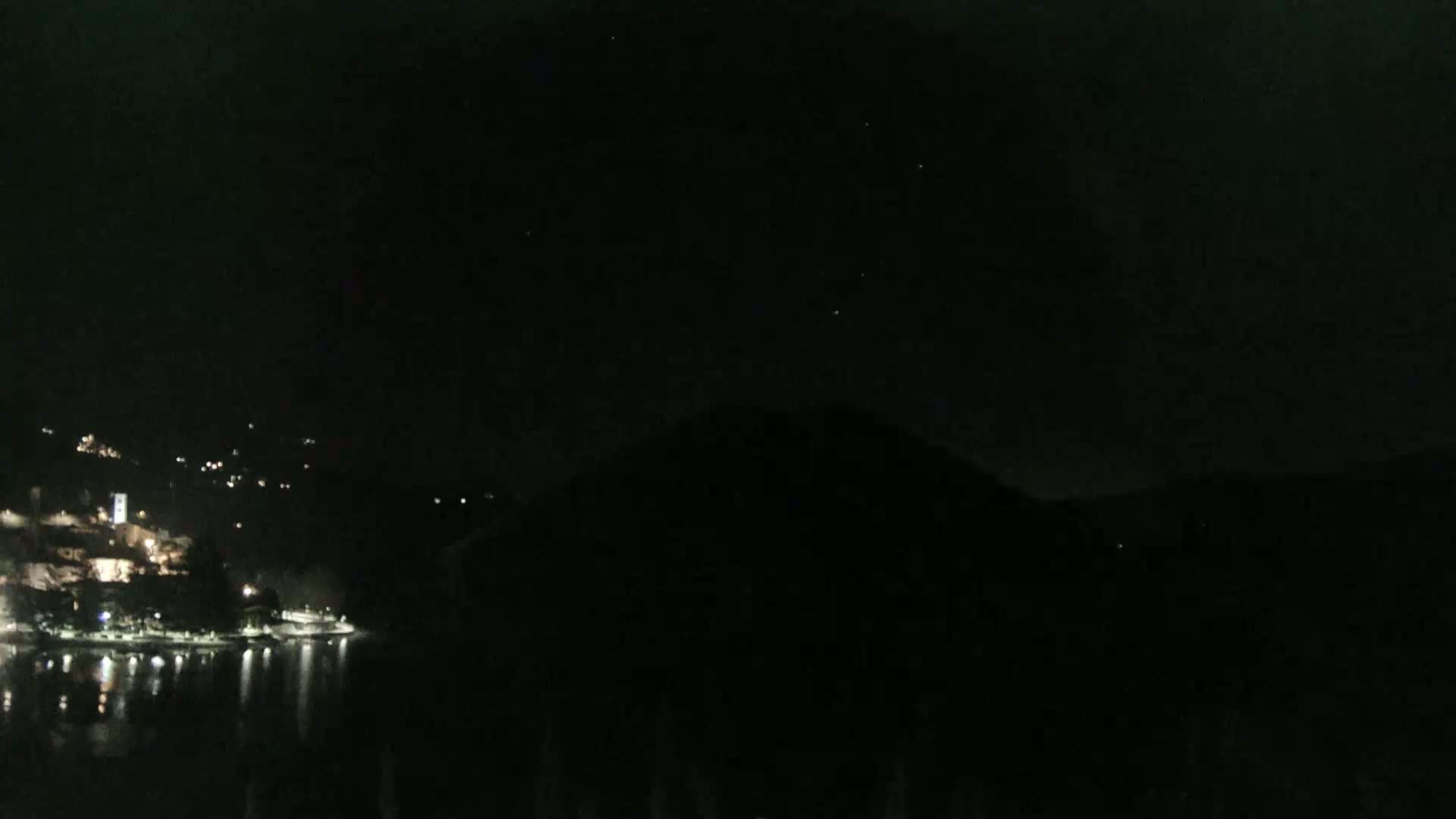 A nighttime view shows a dark, hilly landscape across a body of water reflecting scattered lights from a town on the opposite shore under a clear, starlit sky.