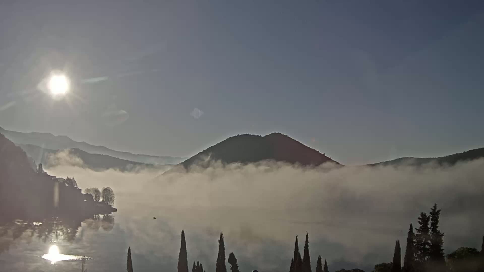 A calm body of water reflects the rising sun, while low-lying fog shrouds the surrounding hills and mountains under a clear sky.