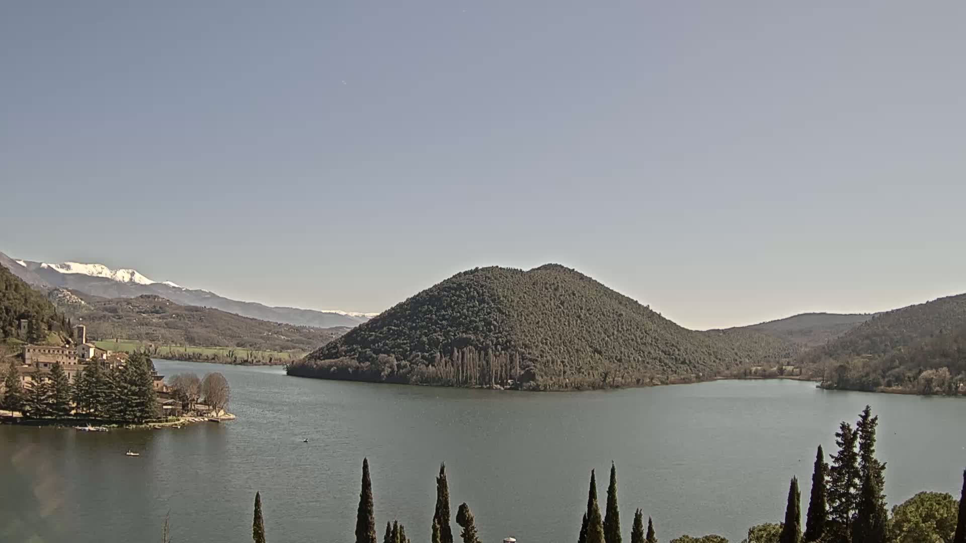 A lake is seen with a wooded island in the middle, a village on the left shore, and snow-capped mountains in the background under a clear sky.