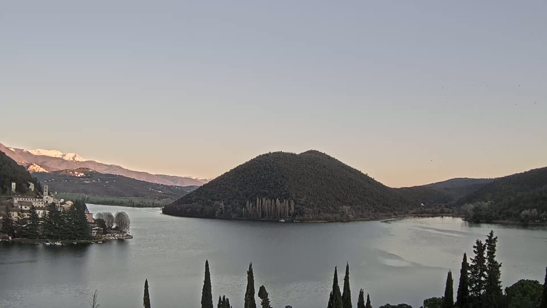 A calm lake is nestled between hills and mountains, one of which has a snow-capped peak, under a clear, pale sky.
