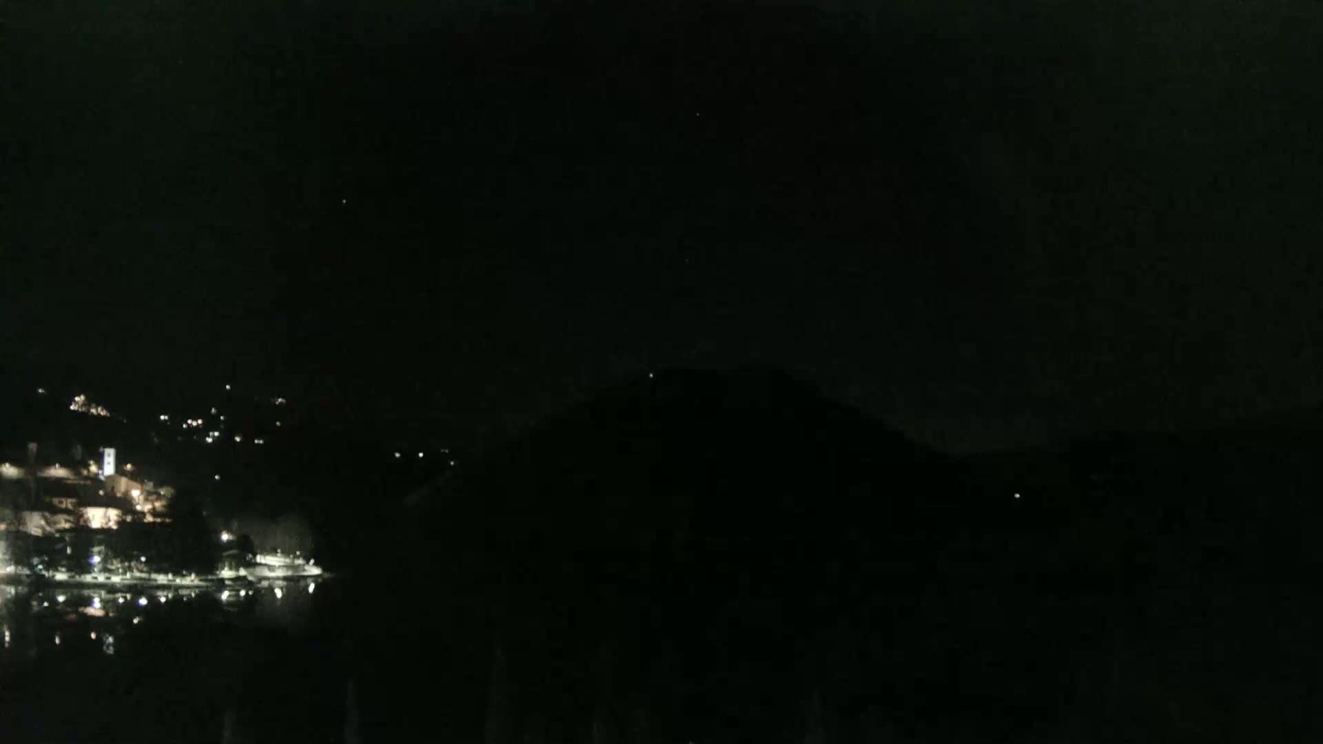 A dark, nighttime scene shows a body of water reflecting the faint lights of a town on the opposite shore, with a dark hill or mountain looming in the foreground under a clear, star-studded sky.