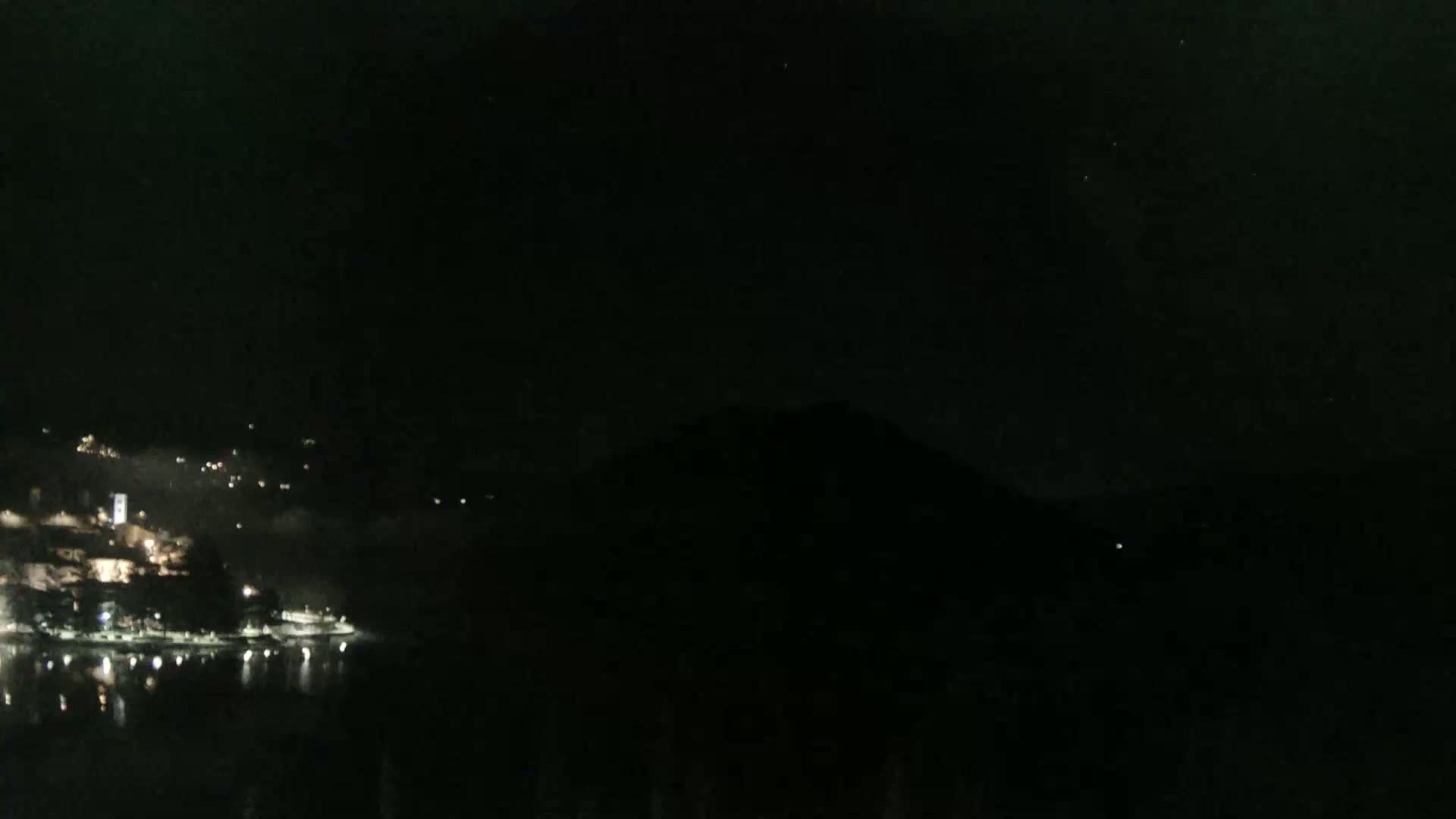 A dark nighttime scene shows a body of water reflecting the lights of a town nestled against a hillside, all under a clear, starlit sky.