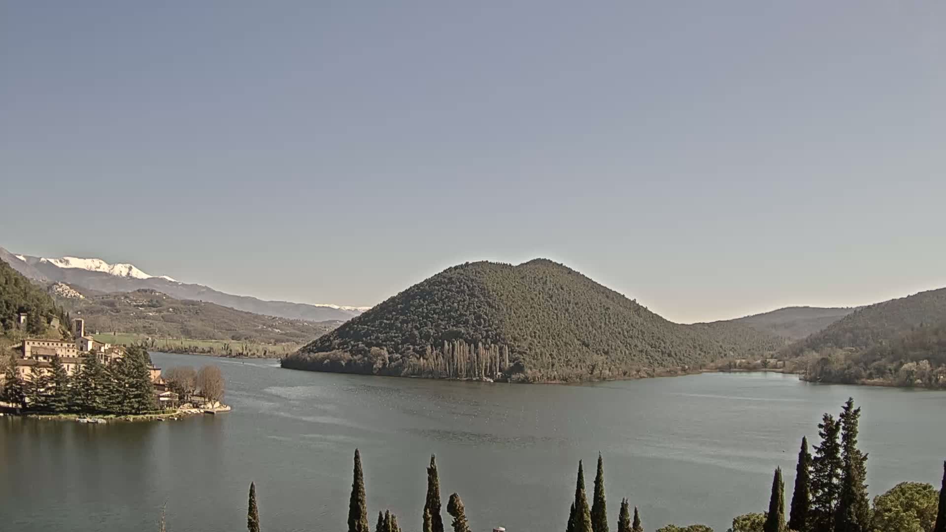 A lake is surrounded by hills and a village, with snow-capped mountains visible in the background under a clear sky.