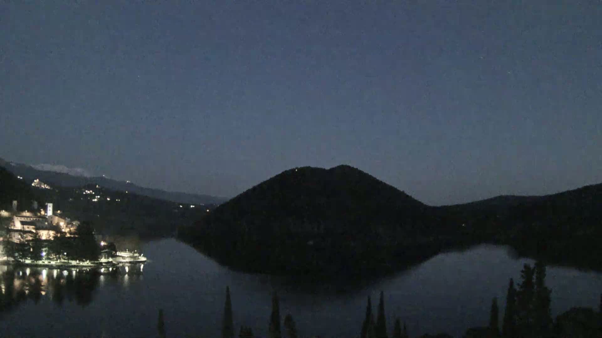 A dark, nighttime scene shows a lake reflecting a village's lights on one side, with a large, dark hill rising from the lake on the opposite side, under a clear, dusky sky.