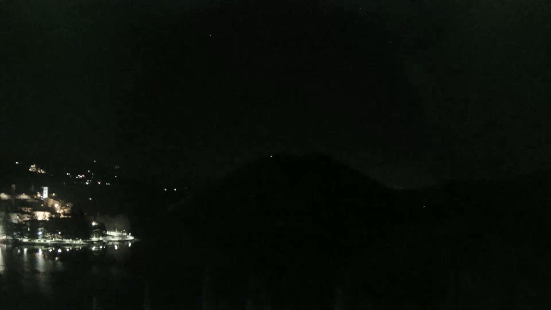 A dark, nighttime view of a town's lights along a body of water, with a dark hill or mountain in the background.