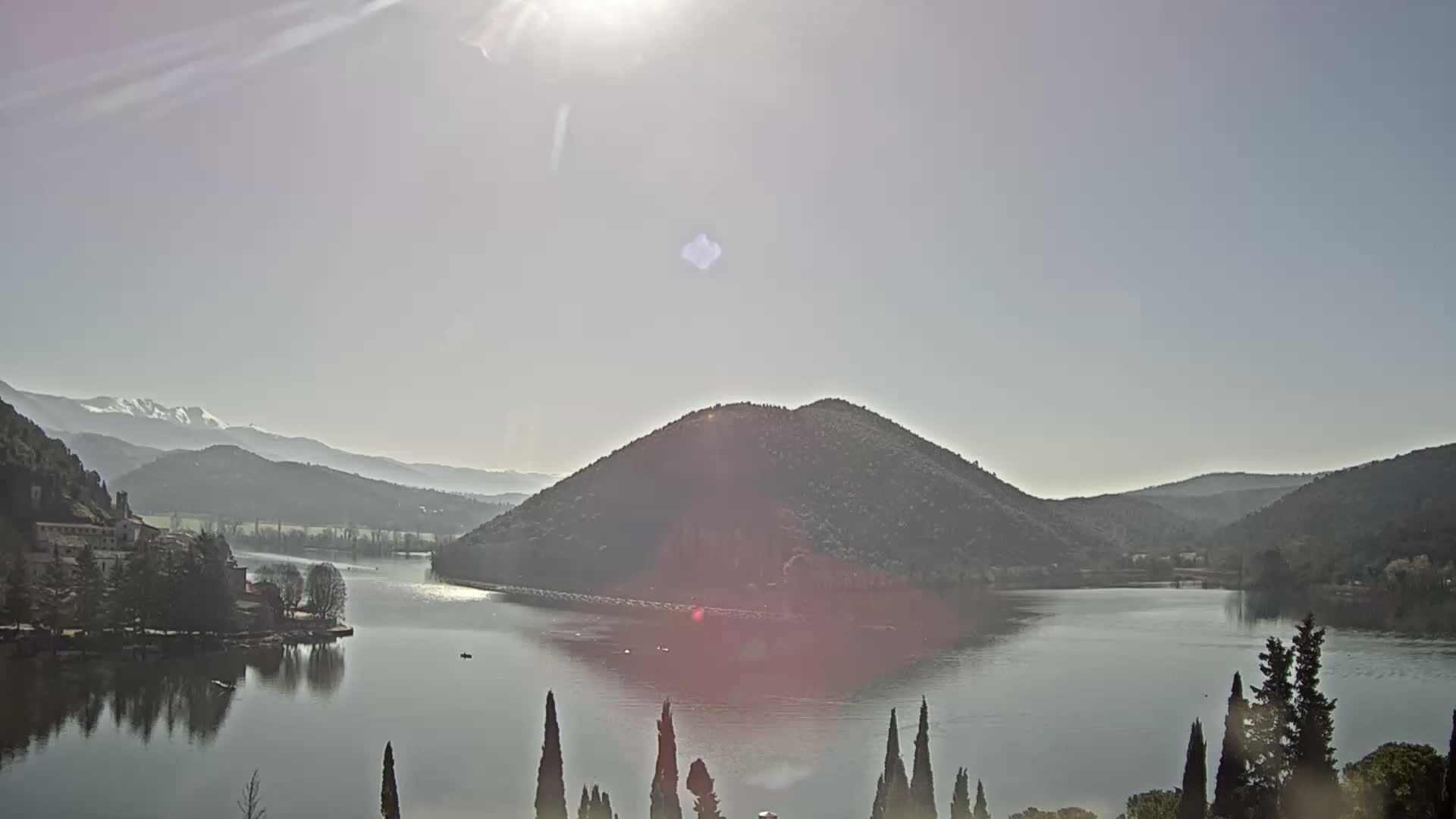A calm lake is surrounded by hills and mountains under a bright, sunny sky, with snow visible on distant peaks.