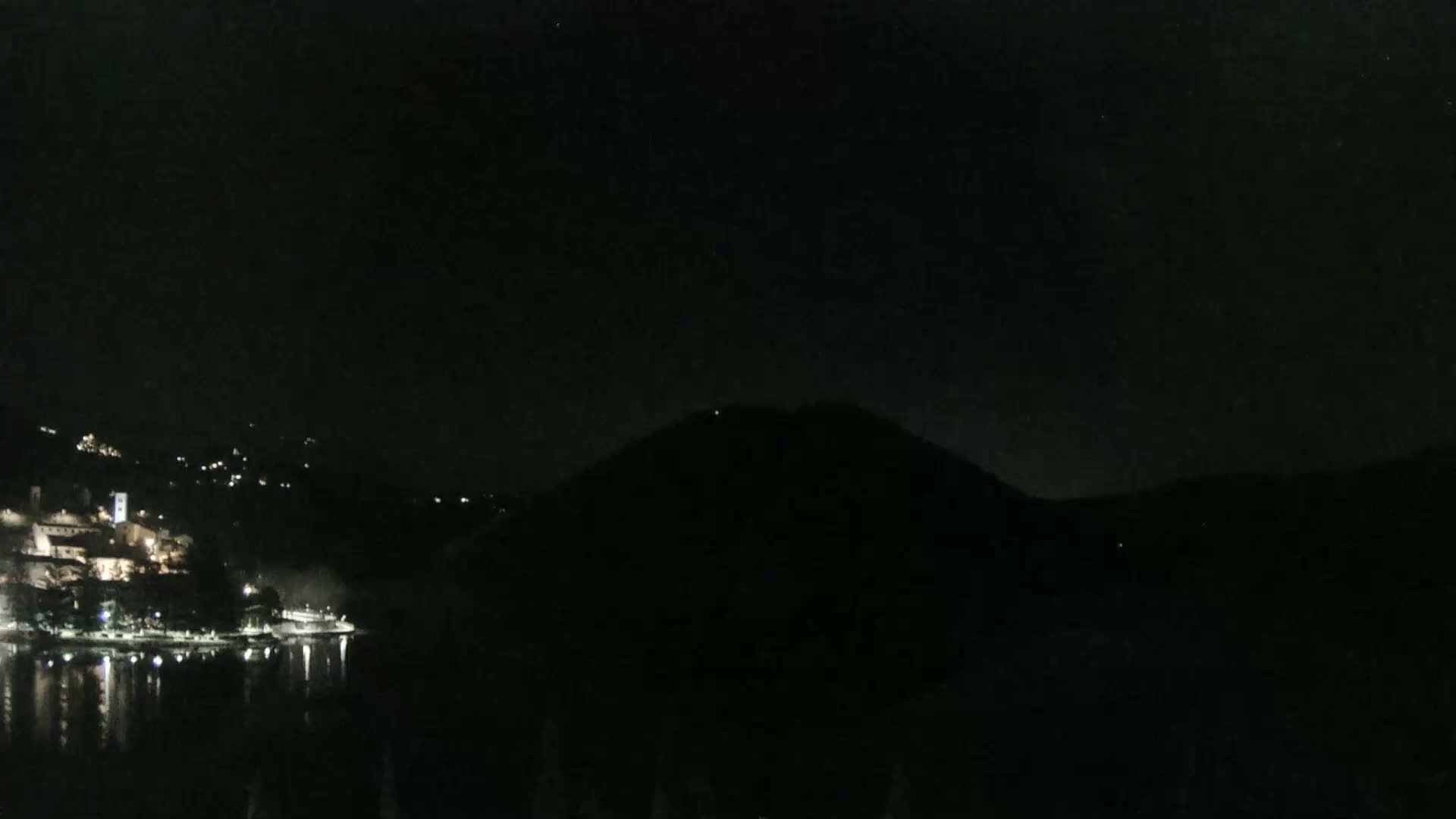 A dark, nighttime scene shows a body of water reflecting the lights of a town nestled at the base of a hill.