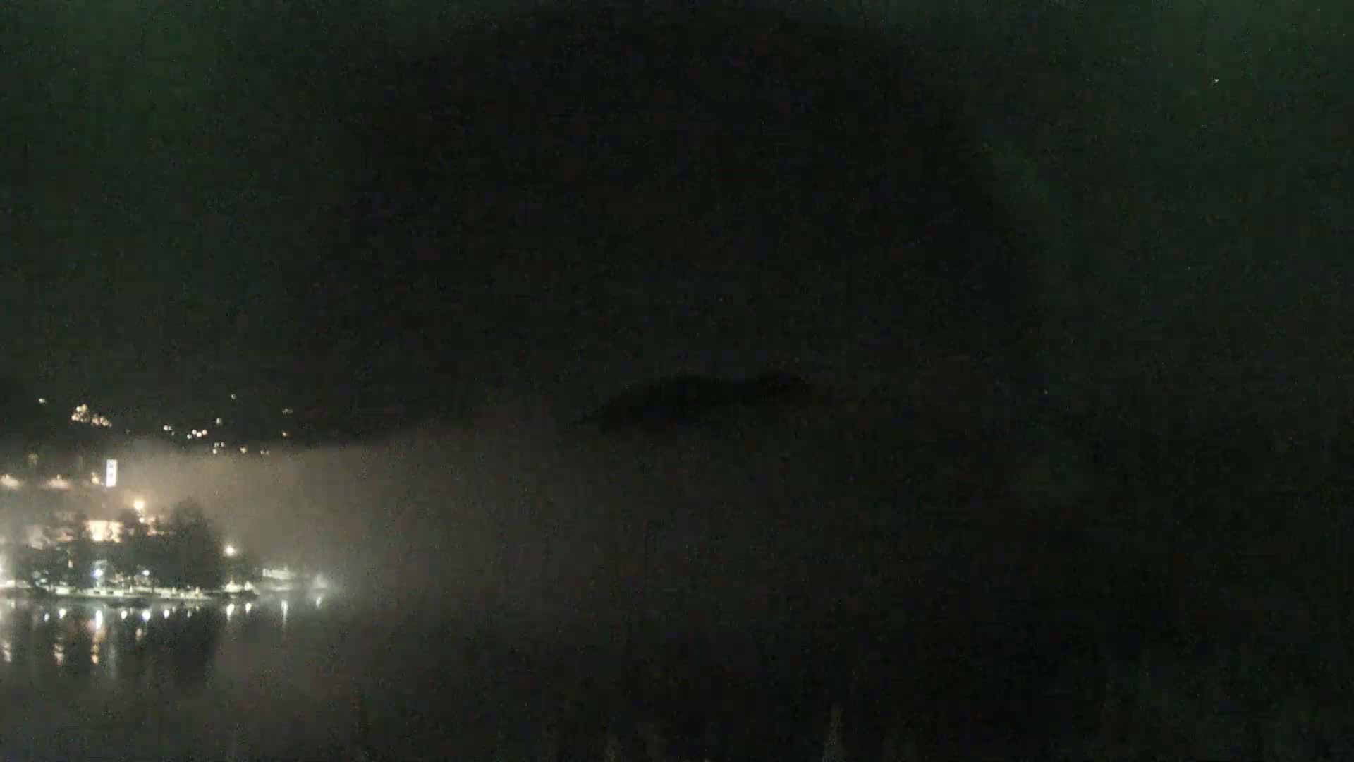 A nighttime view of a town partially obscured by fog along a body of water under a dark sky.