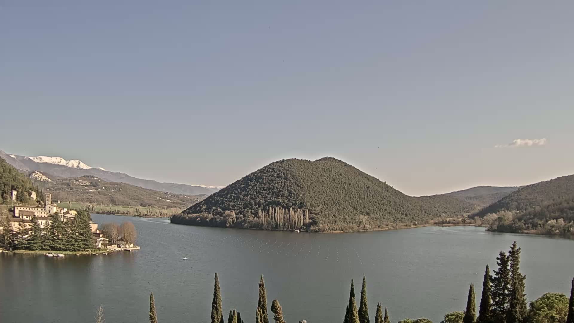 A calm lake is surrounded by hills and mountains, some of which are capped with snow, under a clear sky.
