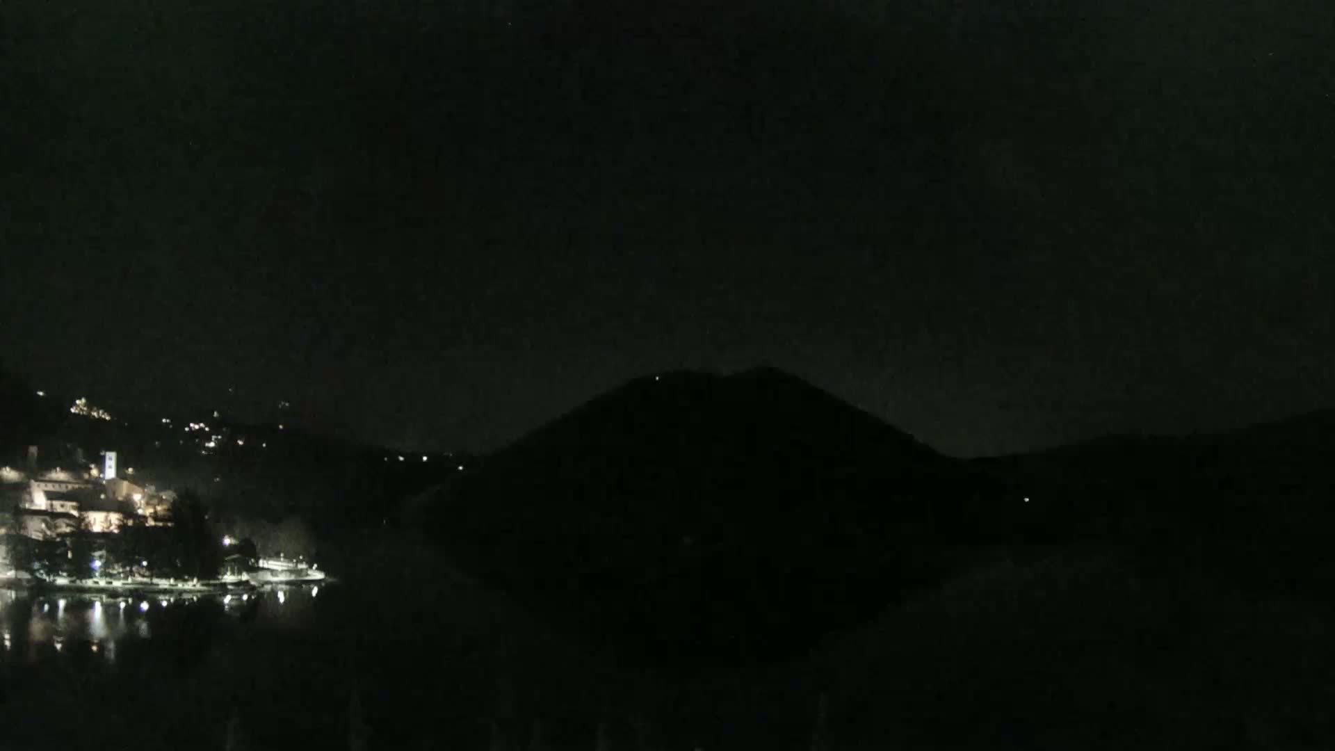 A dark, nighttime view shows a lake reflecting the lights of a town nestled in hills, with a large, dark hill or mountain dominating the center of the image.