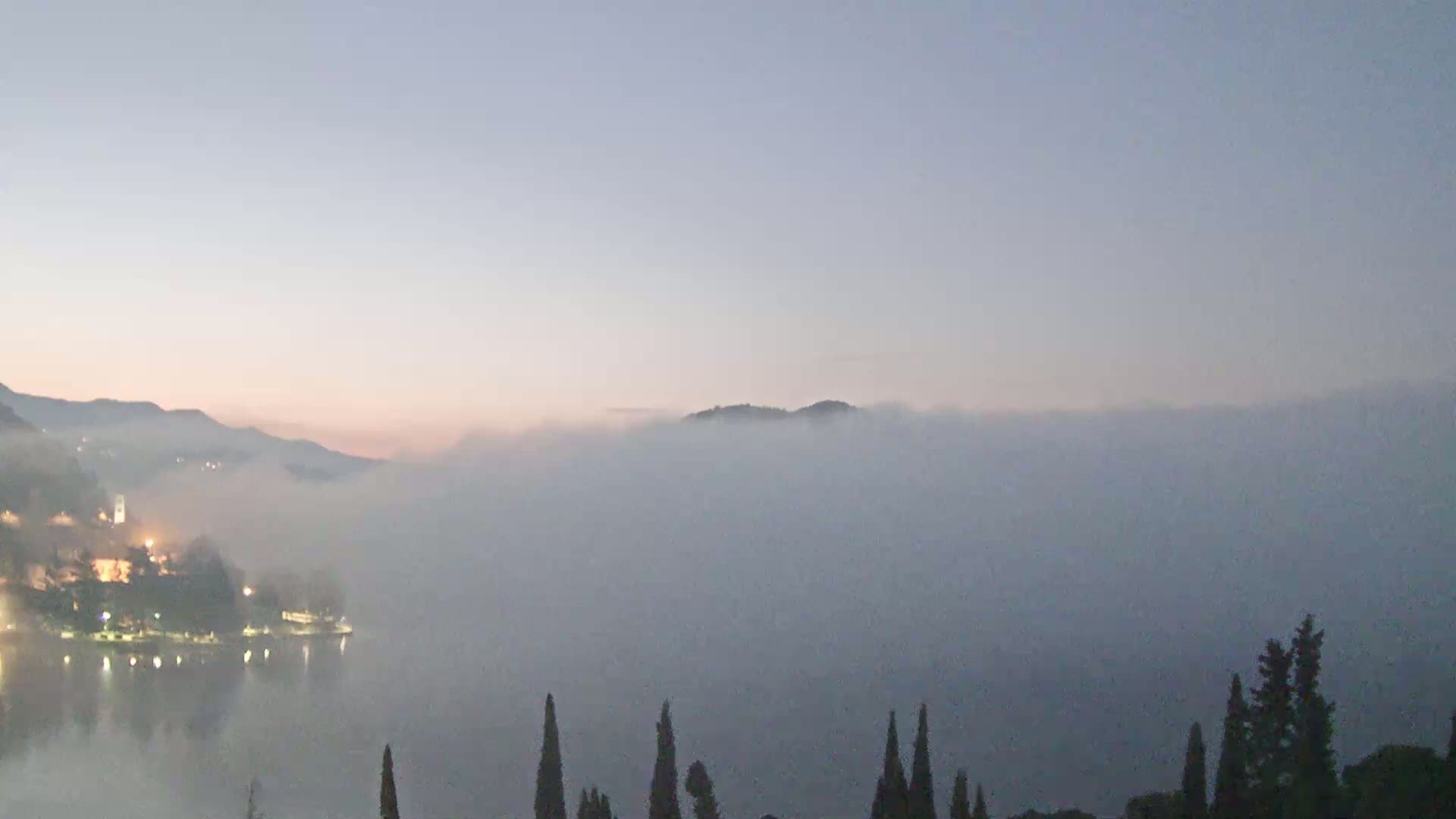 A foggy lake at dawn is nestled between hills, with a town's lights visible along the shore.