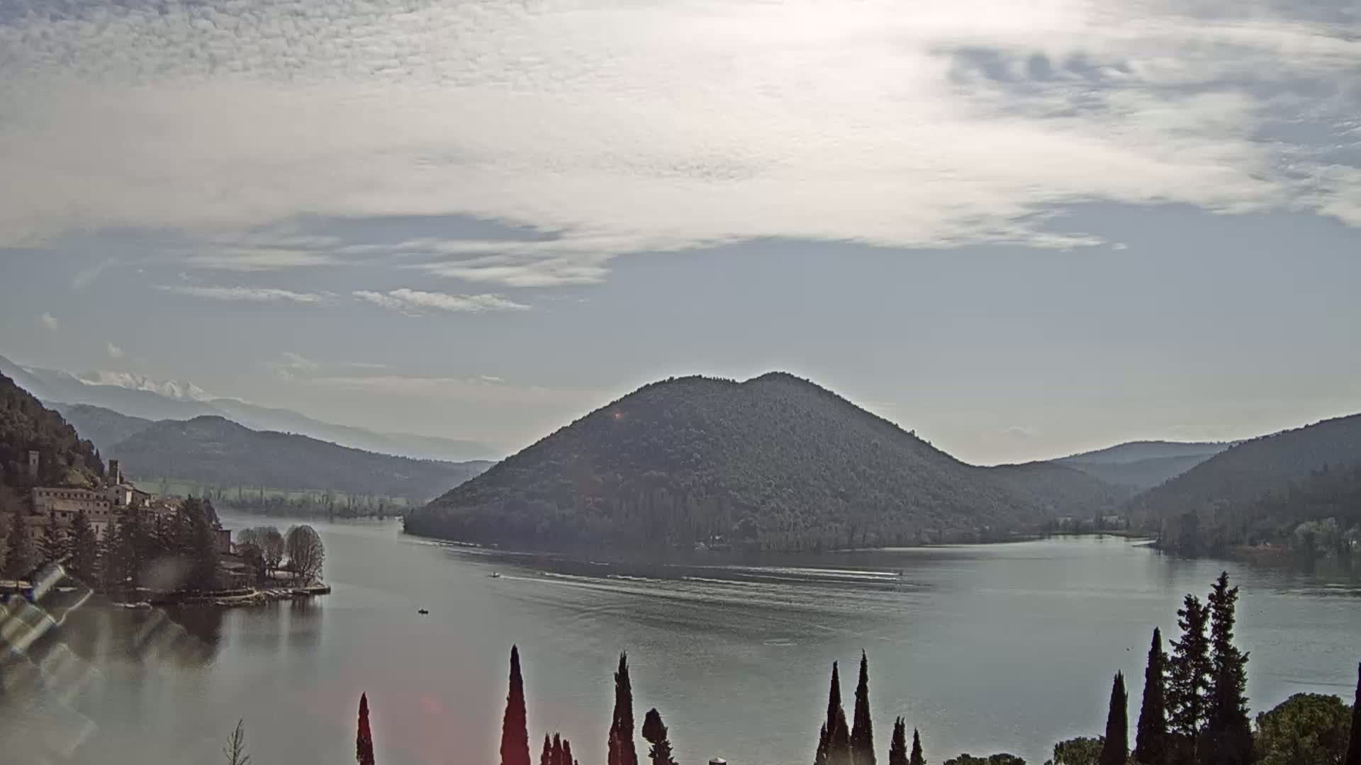 A calm lake is surrounded by mountains and trees under a mostly cloudy sky.