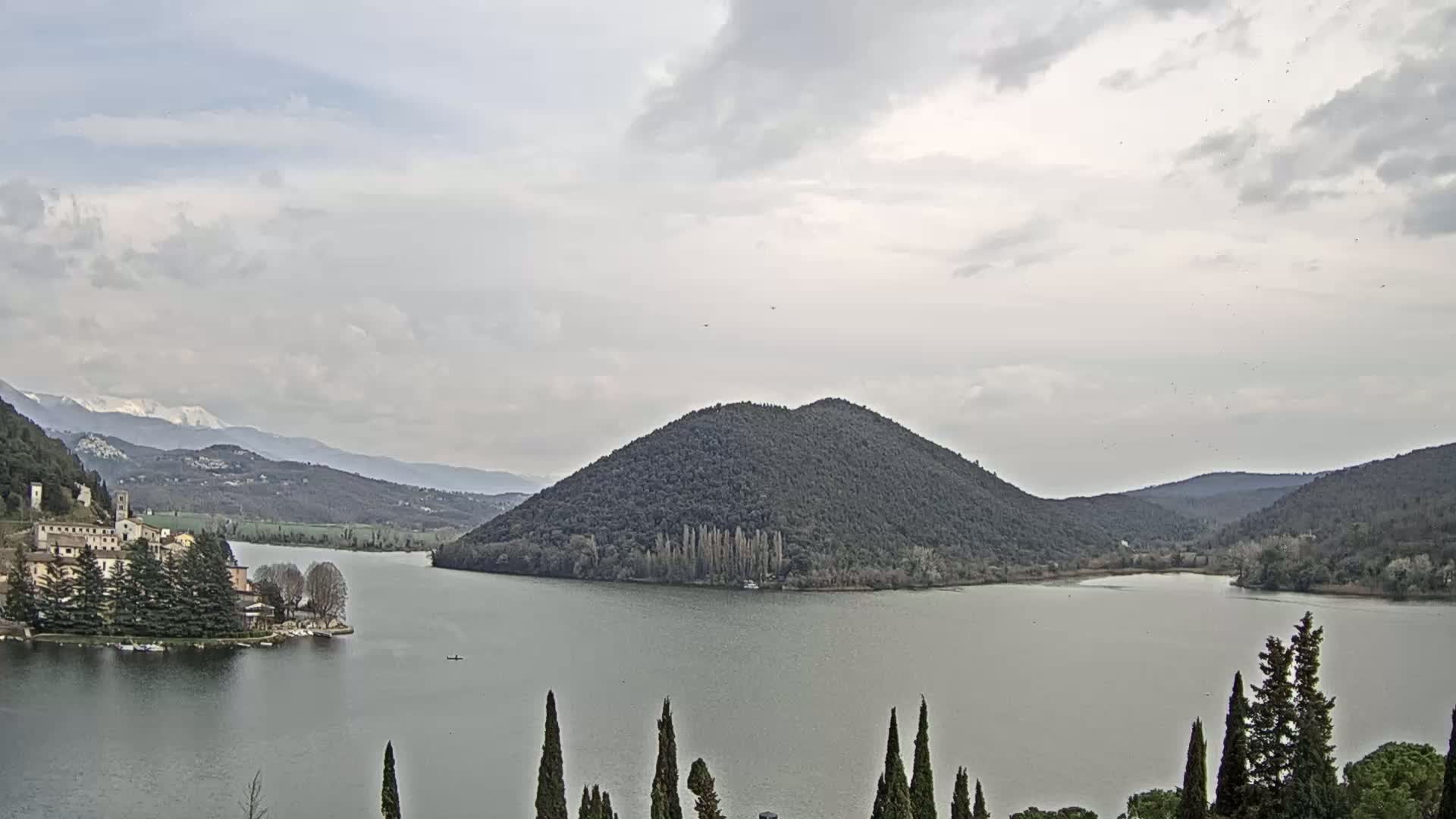 A lake is seen under an overcast sky, with a small village and a tree-covered hill visible in the distance, and snow-capped mountains in the far background.
