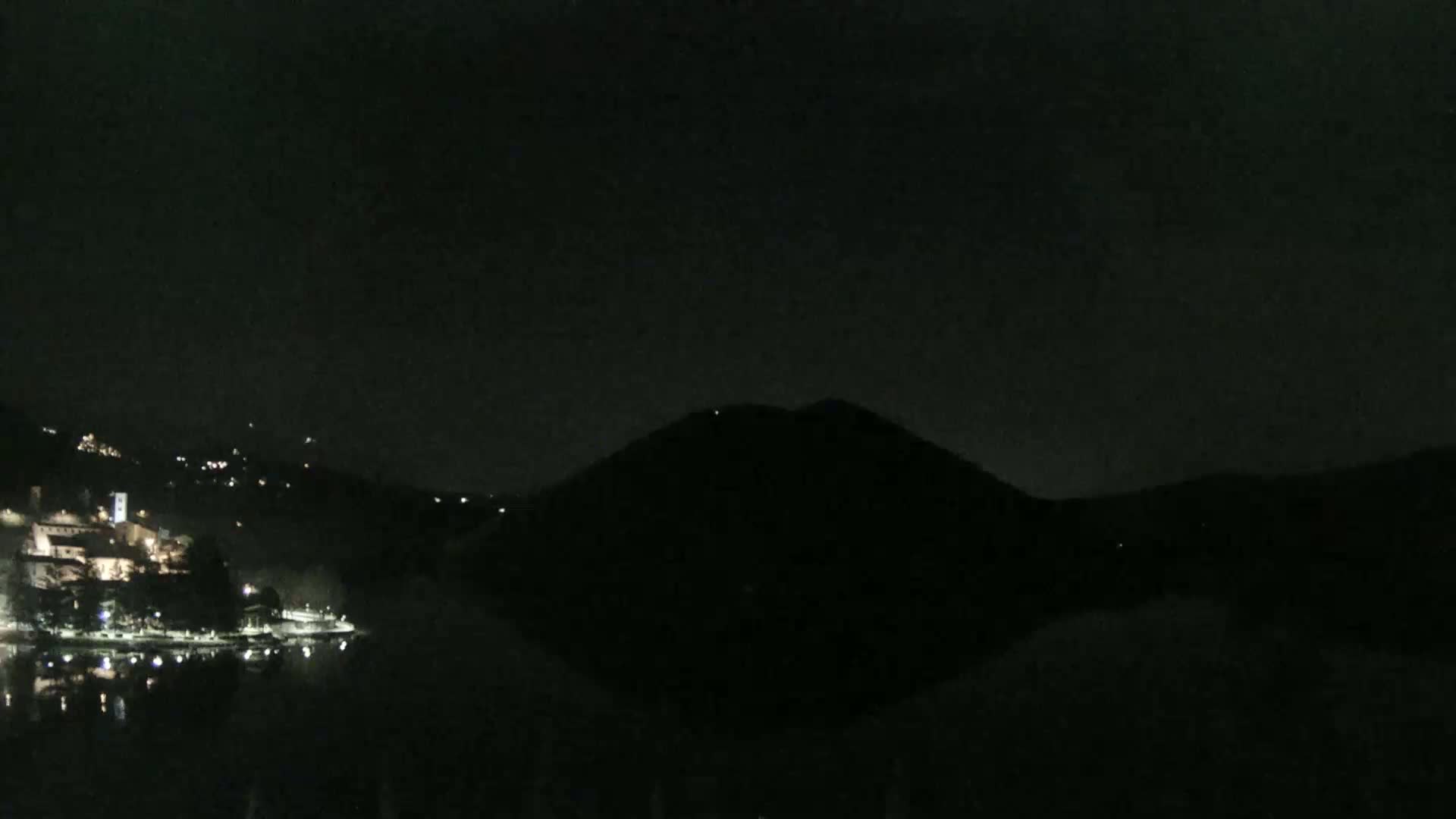 A nighttime view of a dark, hilly landscape with a body of water reflecting the lights of a town on its shore.