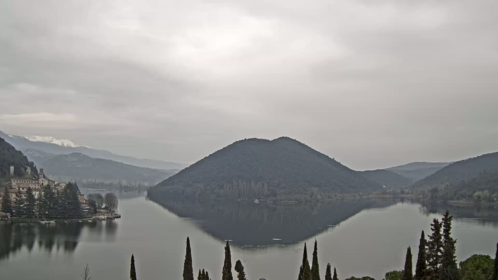 A calm lake reflects a tree-covered hill and distant mountains under an overcast sky.