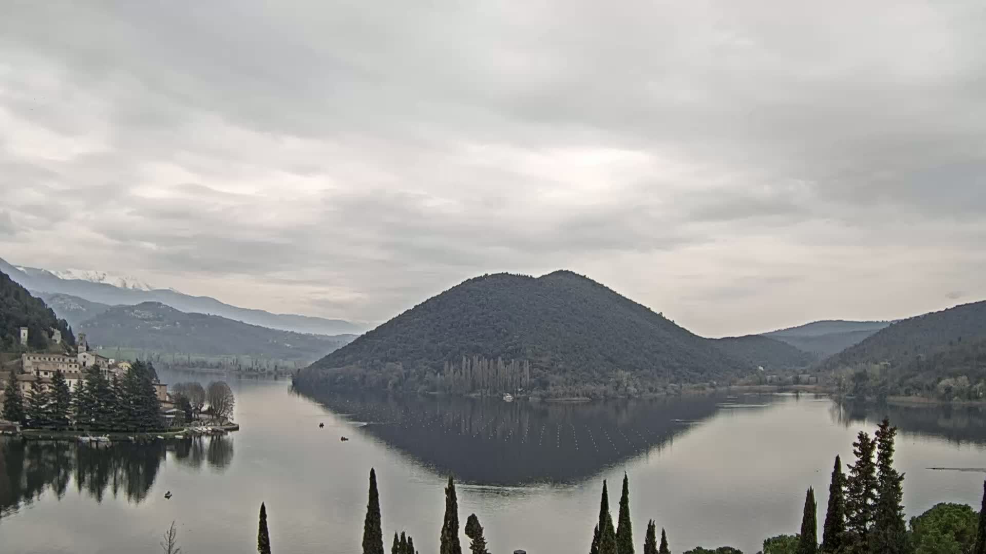 A calm lake reflects a tree-covered hill and snow-capped mountains under a cloudy sky.