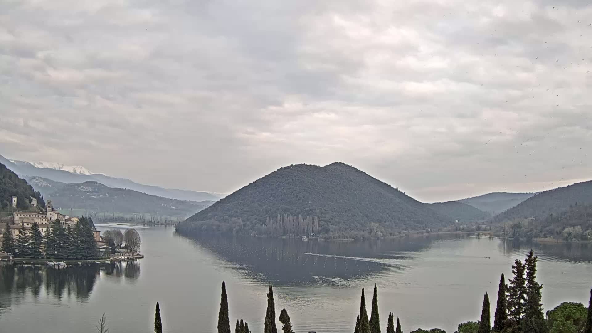 A lake is surrounded by hills and mountains under an overcast sky, with a village visible on the left shore.
