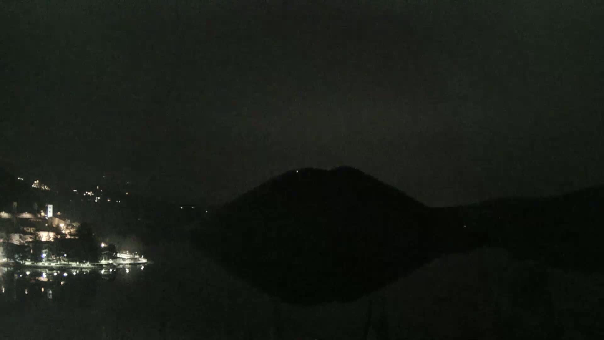 A dark, nighttime scene shows a body of water reflecting the faint lights of a town nestled beside a hill.