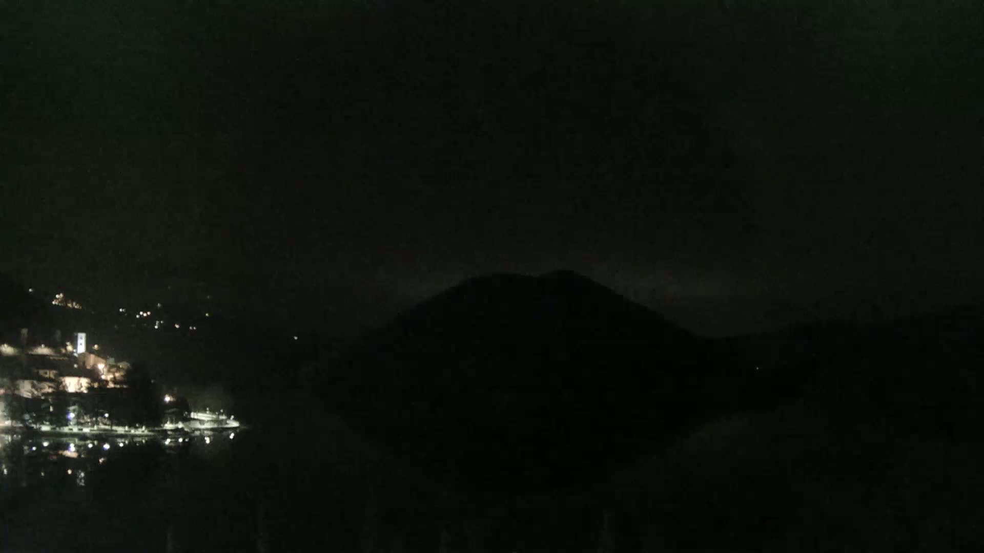 A dark, nighttime view shows a body of water reflecting lights from a town on one side and a large, dark hill on the other.