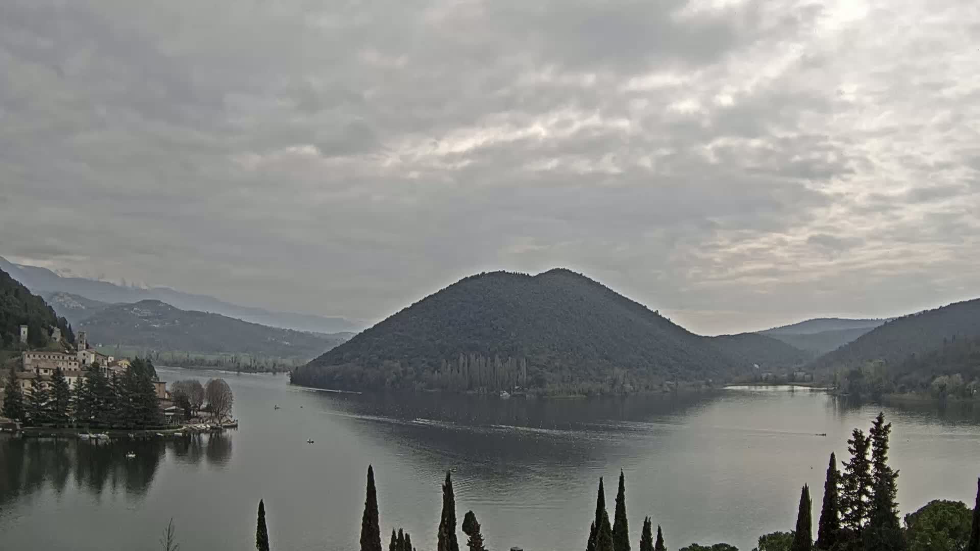 A tranquil lake is nestled between rolling hills under a cloudy sky, with a small village visible on the lake's edge.