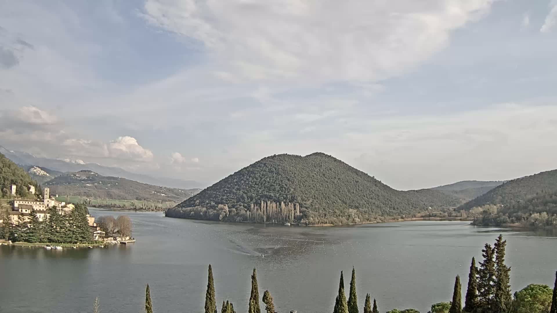 A calm lake is nestled between tree-covered hills under a partly cloudy sky, with a village visible on the lakeshore.